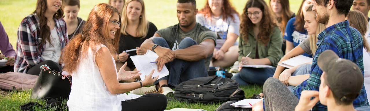 Dr. Val Peterson sitting on the lawn with students for class, smiling.